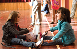 Two primary age children are sitting in a gymnasium floor, rolling a ball between them