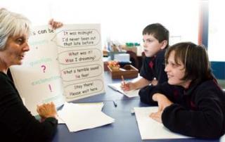 A teacher guiding two primary age students in a reading and writing exercise, at a table.